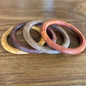 May include: Four wooden bangles in varying shades of brown, tan, and reddish-orange are arranged on a wooden surface. The bangles are circular, with a smooth, polished finish. The wood grain is visible, adding to the natural aesthetic of the jewelry.