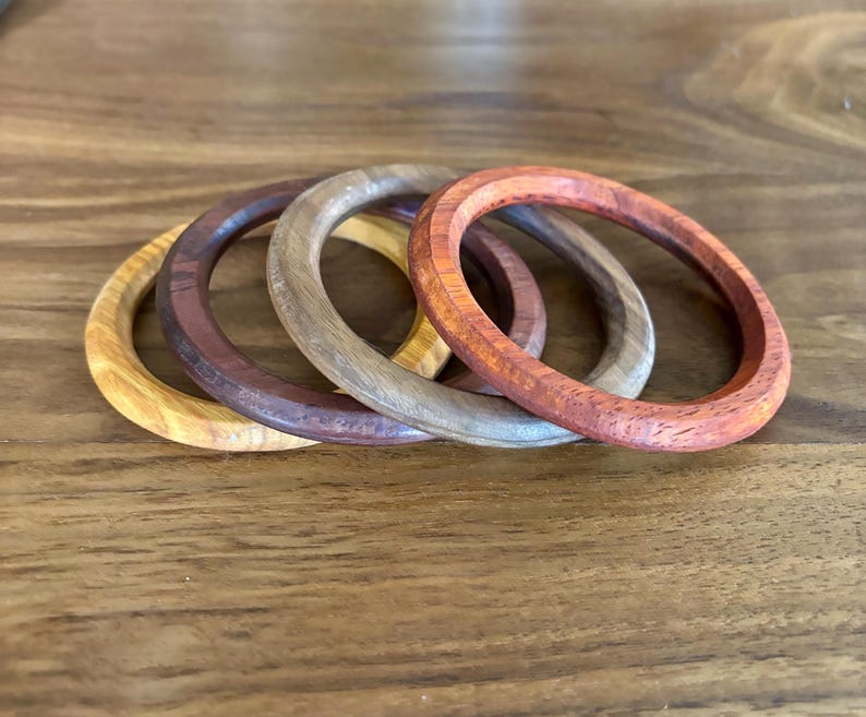 May include: Four wooden bangles in varying shades of brown, tan, and reddish-orange are arranged on a wooden surface. The bangles are circular, with a smooth, polished finish. The wood grain is visible, adding to the natural aesthetic of the jewelry.