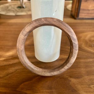 May include: A round, wooden bracelet with a natural wood grain. The bracelet is resting on a wooden surface, with a white vase in the background. The bracelet has a diameter of approximately 3 inches.