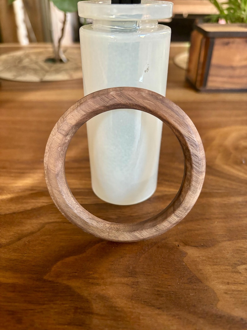 May include: A round, wooden bracelet with a natural wood grain. The bracelet is resting on a wooden surface, with a white vase in the background. The bracelet has a diameter of approximately 3 inches.