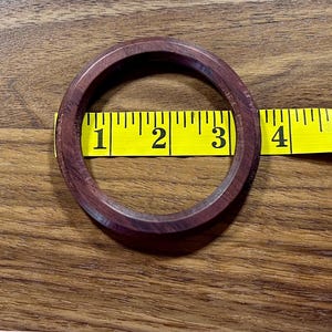 May include: A dark reddish-brown wooden bangle bracelet, approximately 3 inches in diameter, is shown on a wooden surface. A yellow measuring tape is placed next to the bracelet.