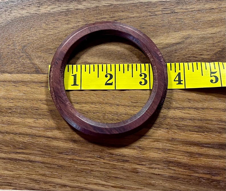 May include: A dark reddish-brown wooden bangle bracelet, approximately 3 inches in diameter, is shown on a wooden surface. A yellow measuring tape is placed next to the bracelet.