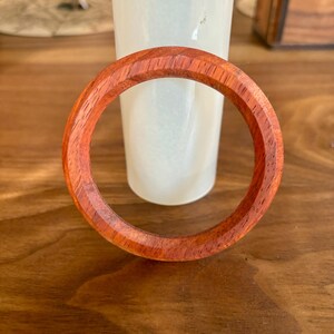 May include: A handcrafted wooden bracelet with a rich reddish-brown hue. The bracelet is circular, with a smooth, polished finish. The wood grain is visible, adding to its natural appeal. The bracelet is approximately 3 inches in diameter.