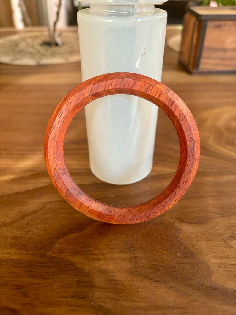 May include: A handcrafted wooden bracelet with a rich reddish-brown hue. The bracelet is circular, with a smooth, polished finish. The wood grain is visible, adding to its natural appeal. The bracelet is approximately 3 inches in diameter.