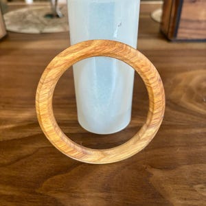 May include: A light brown wooden bangle bracelet with a circular design. The bracelet is made of wood and has a natural grain pattern. A light blue vase is in the background.