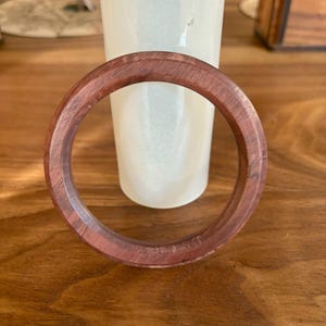 May include: A round, reddish-brown wooden bracelet. The bracelet is made of a solid piece of wood and has a smooth, polished finish. The bracelet is approximately 3 inches in diameter.