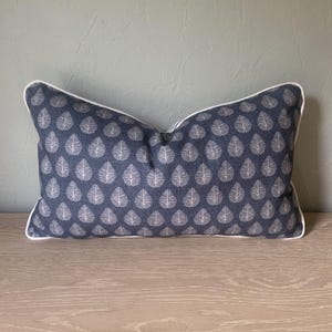 May include: A rectangular throw cushion featuring a dark blue fabric base with a repeating white leaf pattern. The cushion is finished with a crisp white piping trim along all four edges, providing a clean and defined border to the overall design.