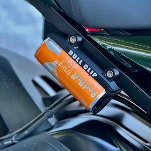 Könnte beinhalten: Ein schwarzer "Bull Clip" hält eine orangefarbene Red Bull-Dose an einem Motorrad. Die Dose hat einen silbernen Deckel und den Text "The AP Edition". Der Clip ist in der Nähe des Sitzes montiert.