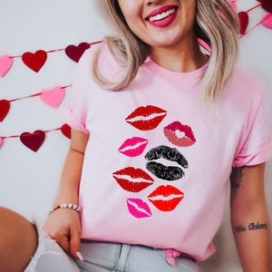 以下が含まれることがあります： 赤、ピンク、黒のリッププリントのデザインが施された、明るいピンク色のTシャツ。半袖のクルーネックです。バレンタインデーをテーマにしています。
