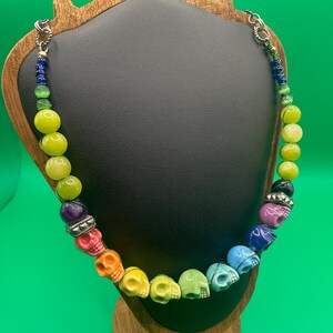 Op de afbeelding: Een kleurrijke ketting met schedelvormige kralen in een regenboog van kleuren, waaronder roze, oranje, geel, groen, blauw en paars. De ketting bevat ook groene en gele ronde kralen, geregen aan een zilveren ketting.