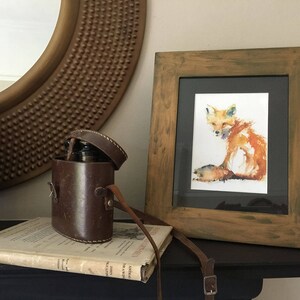 May include: A watercolour painting of a fox in a wooden frame. The fox is sitting on a white background and is painted in shades of orange, red, and yellow. The frame is a dark brown wood with a distressed finish.