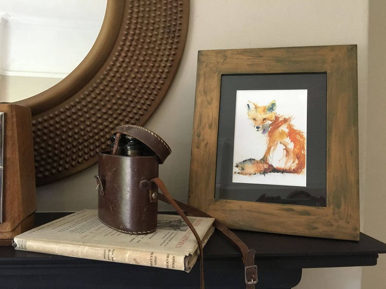 May include: A watercolour painting of a fox in a wooden frame. The fox is sitting on a white background and is painted in shades of orange, red, and yellow. The frame is a dark brown wood with a distressed finish.