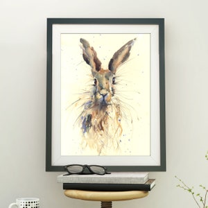 May include: Watercolor painting of a hare in a black frame with a white mat. The hare is depicted with brown and tan fur, long ears, and detailed whiskers. Blue paint splatters accent the background. The framed artwork is displayed on a wall.