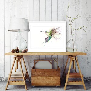 May include: A watercolour painting of a hummingbird in flight, framed and displayed on a wooden shelf with a lamp and vase.