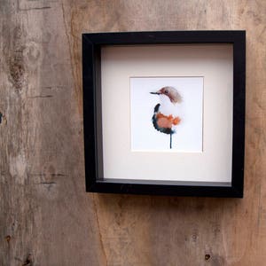 May include: A watercolour painting of a bird in a black frame. The bird is brown, white, and blue, and is perched on a branch. The painting is on a white background.