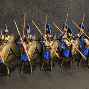 May include: A collection of miniature Spartan warriors, each with a spear and shield. The figures wear gold armour, blue tunics, and helmets, with shields marked with the letter "A". The spears are wooden, and the figures are arranged in a battle formation.