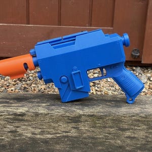 May include: A blue toy blaster with a bright orange muzzle attachment. The blaster features a blocky, angular design with a textured grip, a trigger, and a rectangular magazine well. The construction appears to be made of plastic with visible layer lines from 3D printing, showcasing a solid, monochromatic blue body with a contrasting orange front piece.