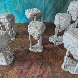 Peut inclure: Plusieurs structures grises en forme de rochers avec des surfaces texturées, soutenues par de fins piliers verticaux. Elles sont disposées sur une surface brune, avec une toile de fond bleue et verte. Les structures varient en taille et en forme.