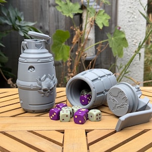 May include: A grey 3D-printed grenade-shaped container with a skull emblem, featuring a removable top lid. Beside it, a second grenade container lies on its side, revealing several purple and cream-colored polyhedral dice scattered nearby.