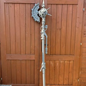 Può includere: Un'ascia fantasy alta e grigia con un design a teschio e ingranaggi. L'ascia presenta un lungo palo con dettagli intricati, tra cui tubi ed elementi meccanici. La testa dell'ascia ha un design a lama di sega circolare e un teschio in cima.