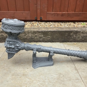 May include: A gray, intricately designed hammer with a textured handle and decorative head. The hammer is resting on a gray stand, showcasing its detailed craftsmanship. The hammer's head features a circular design with a geometric pattern.
