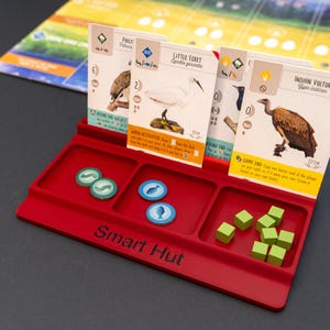 May include: A red plastic game organiser with three compartments, holding game cards, green and blue tokens, and green wooden cubes. The cards feature bird illustrations and text. The organiser has the text "Smart Hut" on the front.