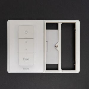 May include: A white Philips Hue smart light switch with three buttons and the brand name "hue" printed on the front. The switch is mounted on a white frame, with a black background.