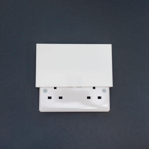 May include: A white double electrical socket with a hinged cover. The socket has two sets of three-pin sockets. The cover is rectangular and covers the sockets. The background is a dark grey.