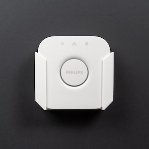 May include: A white Philips Hue smart home hub with a round button in the centre and three small buttons on the top. The hub is mounted on a white wall mount.