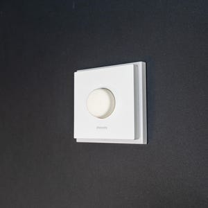 May include: A white light dimmer switch with a round knob, mounted on a dark grey wall. The switch has a square faceplate with the brand name "Philips" printed below the knob.