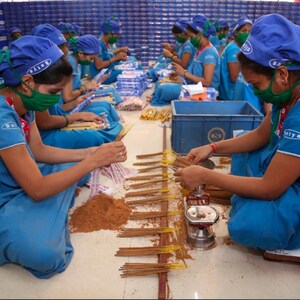 K&ouml;nnte beinhalten: Eine Gruppe von Menschen in blauen Uniformen und Kopfbedeckungen arbeitet an einem Produktionsband und montiert R&auml;ucherst&auml;bchen. Sie sitzen auf dem Boden und verwenden ihre H&auml;nde, um die St&auml;bchen zusammenzubauen. In der Mitte des Bildes befindet sich ein Stapel R&auml;ucherst&auml;bchen.
