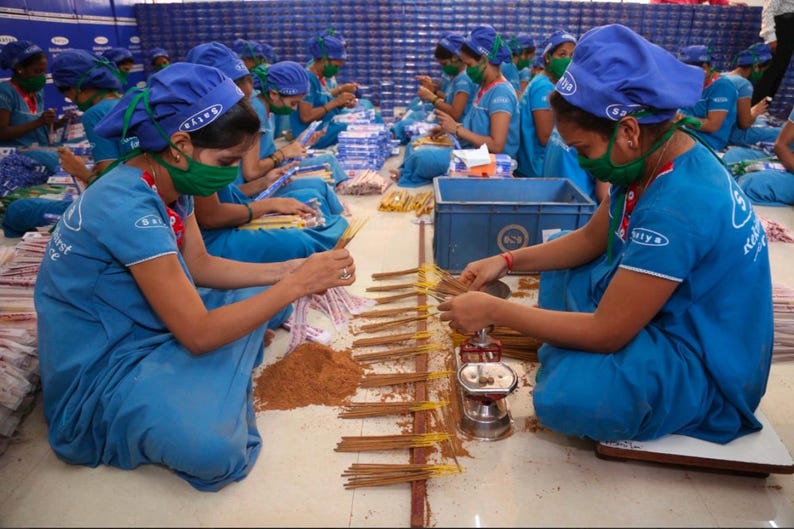 K&ouml;nnte beinhalten: Eine Gruppe von Menschen in blauen Uniformen und Kopfbedeckungen arbeitet an einem Produktionsband und montiert R&auml;ucherst&auml;bchen. Sie sitzen auf dem Boden und verwenden ihre H&auml;nde, um die St&auml;bchen zusammenzubauen. In der Mitte des Bildes befindet sich ein Stapel R&auml;ucherst&auml;bchen.