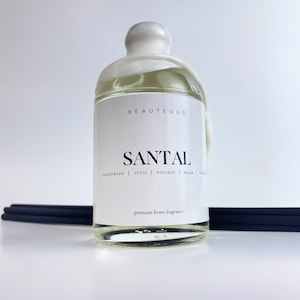 Peut inclure: Une bouteille en verre transparent avec un bouchon blanc et une étiquette blanche sur laquelle est inscrit "BEAUTEOUS SANTAL sandalwood | orris | coconut | musk | amber premium home fragrance".