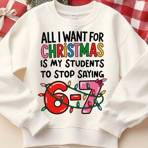 Könnte beinhalten: Cremefarbenes Sweatshirt mit dem Text "ALL I WANT FOR CHRISTMAS IS MY STUDENTS TO STOP SAYING 6-7". Die Zahlen 6 und 7 sind mit Lichterketten verziert. Eine rote Geschenkbox und Weihnachtsdekorationen sind im Hintergrund.