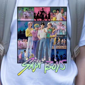 Puede incluir: Camiseta blanca con un gráfico colorido de un grupo de personas y el texto "Saint Boys" en verde neón y amarillo. La camiseta la lleva una persona con una mochila gris.
