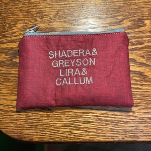 Pode incluir: Uma bolsa bordô com zíper e texto bordado "SHADERA & GREYSON LIRA & CALLUM". A bolsa tem um zíper prateado e é retangular. Sobre uma superfície de madeira.