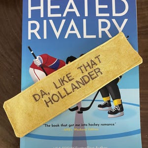Könnte beinhalten: Ein Buch mit dem Titel "HEATED RIVALRY" mit einem gelben Lesezeichen mit der Aufschrift "DA, LIKE THAT HOLLANDER". Das Buchcover zeigt Eishockeyspieler. Der Autor ist Rachel Reid.