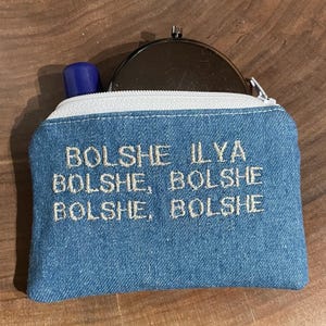 May include: A small, blue denim pouch with a white zipper and the embroidered text "Bolshe Ilya Bolshe, Bolshe Bolshe". A blue lipstick tube and a silver compact mirror are visible inside the pouch.