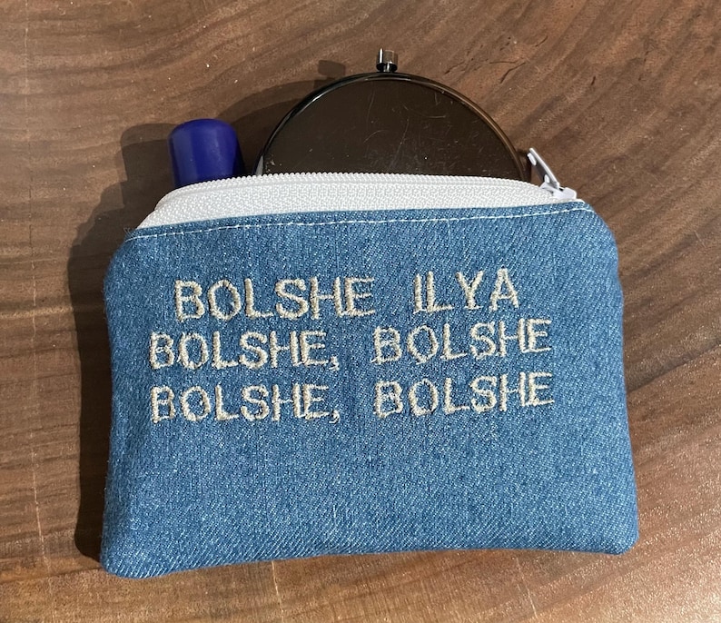 Pu&ograve; includere: Piccola pochette in denim blu con cerniera bianca e la scritta ricamata "Bolshe Ilya Bolshe, Bolshe Bolshe". Un tubetto di rossetto blu e uno specchietto compatto argentato sono visibili.