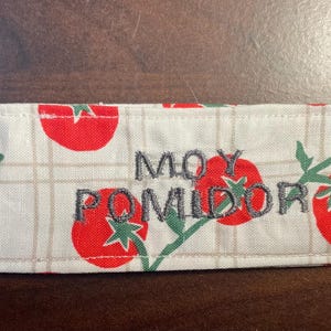 May include: A rectangular fabric item with a white background featuring a repeating pattern of red tomatoes and green stems. The embroidered text "MOY POMIDOR" is in the center. The item has a stitched border.