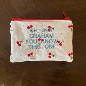 Pode incluir: Uma bolsa de tecido branco com fecho de correr, apresentando um padrão repetitivo de pequenas cerejas vermelhas e pontos pretos minúsculos. O centro está bordado com o texto azul "OH, SHIT GRAHAM YOU KNOW THIS ONE" e tem um fecho vermelho.