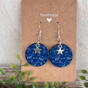 May include: A pair of handmade earrings with a navy blue circular design featuring the letters "FDT" and a bicycle pattern. Each earring has a silver star charm and a silver hook. The earrings are displayed on a brown card.