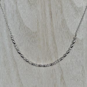 May include: A silver necklace with a delicate chain and a series of oval and cylindrical beads. The beads are evenly spaced along the necklace, creating a minimalist and elegant design. The necklace is displayed on a light wooden surface.