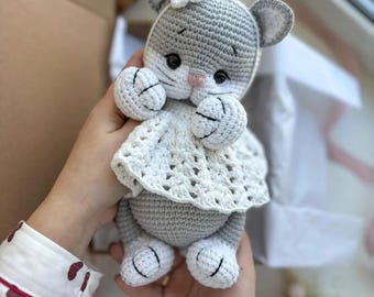 Peut inclure: Un jouet en crochet en forme de chat gris avec une robe blanche et une fleur rose sur la tête.