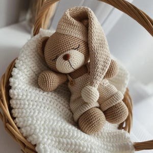 Puede incluir: Un oso de peluche de crochet con un gorro de dormir y un mono de crochet. El oso está acostado en una cesta de crochet blanca.