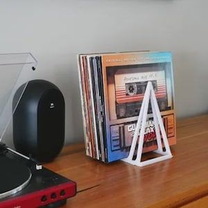 May include: A white, triangular vinyl record holder supporting a stack of vinyl records. The album cover visible features a cassette tape design with the text "Awesome Mix Vol. 2". A turntable and speaker are also visible.