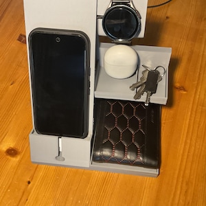 May include: A light gray multi-functional desk organizer stand featuring a dedicated slot for a smartphone with a charging cable, a raised platform for a smartwatch, a small tray for earbuds, a key hook, and a lower compartment for a wallet.