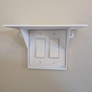 Op de afbeelding: Wit plasticen plank met twee lichtschakelaars eronder gemonteerd. De plank heeft een kleine lip aan de voorkant en twee kleine haken aan de zijkanten.
