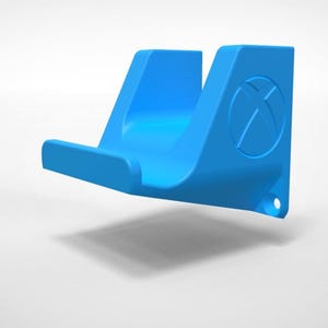 Puede incluir: Un soporte de consola de juegos azul brillante para montar en la pared. El soporte presenta un diseño curvo con un logotipo de Xbox en el lateral. Tiene una estructura de gancho para sujetar la consola de forma segura. El soporte es de material liso.