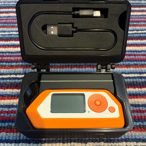 May include: A black carrying case with a small orange and white device inside. The device has a screen and buttons. A black USB cable is included.